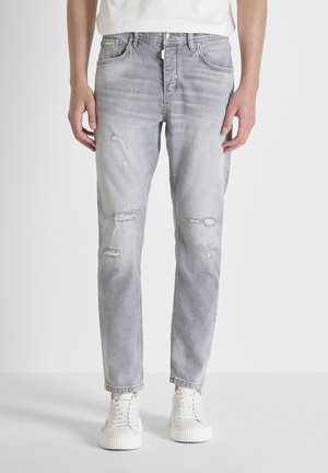 Hombre vistiendo jeans grises claros desgastados y zapatillas blancas contra un fondo blanco liso.