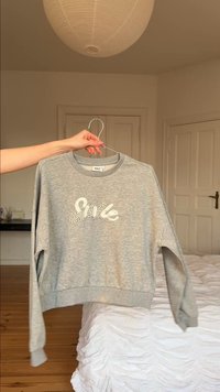 Φωτεινό γκρι φούτερ με το "Smile" σε λευκό, κρατημένο από ένα χέρι σε κρεμάστρα σε ένα υπνοδωμάτιο με κρεβάτι και κλειστές πόρτες.