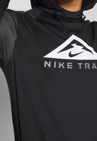 Czarna bluza z kapturem z trójkątnym białym logo z nadrukiem i tekstem "NIKE TRA" w szarym kolorze na klatce piersiowej.