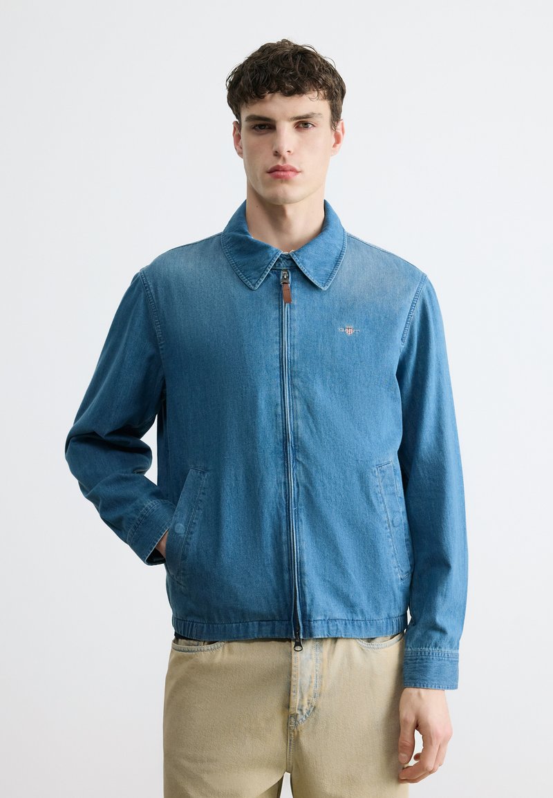 Νεαρός άνδρας που φοράει ένα γαλάζιο τζιν μπουφάν με φερμουάρ και γιακά, καθώς και μπεζ παντελόνι, στέκεται μπροστά σε απλό λευκό φόντο.