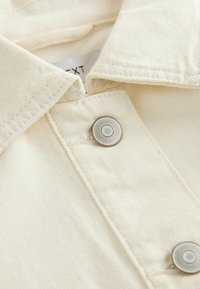 Gros plan sur un tissu de couleur crème avec des coutures visibles, des boutons-pression en métal et une étiquette de vêtement partiellement visible.