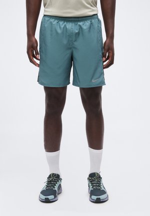 Mann iført turkise Nike treningsshorts, hvite sokker og svarte og turkise løpesko, står mot en ensfarget bakgrunn.