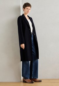 Manteau noir en laine au design ajusté, avec un col cranté, trois boutons et des poches latérales, associé à un jean bleu à jambes larges et des bottes marron.