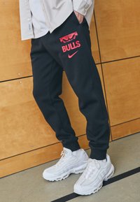 Sorte joggebukser har et rødt grafisk design med "BULLS" og Nike-logo. Kombinert med hvite joggesko, satt mot en trevegg.