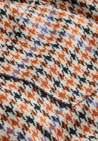 Woven textile featuring an orange, black, and white houndstooth pattern with hints of purple. Smooth texture, visible seams, and a folded edge.