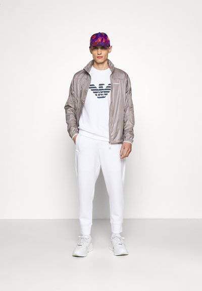 Coupe-vent gris clair, t-shirt avec logo blanc, joggers blancs et baskets argentées. Casquette multicolore avec motif floral. Textures lisses.