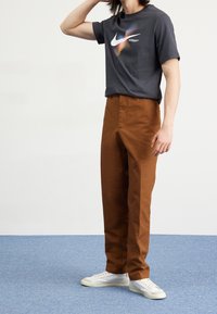 Camiseta gris con el logotipo de Nike multicolor, combinada con pantalones marrones. El modelo lleva zapatillas blancas. Diseño minimalista y ajuste casual.