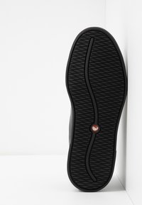 Semelle de chaussure noire avec un motif ondulé et un logo circulaire rouge près du centre, positionnée sur un fond blanc d’angle.