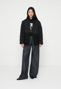 Melnā pūkaina jaka ar pogu aizdari un priekšējiem kabatām, virs melna kapuces. Modelis ģērbjas plašās džinsās bikses un melnās kurpes.