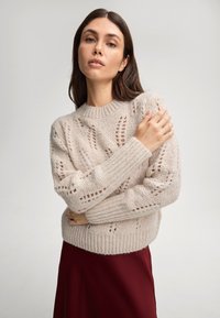 Hellbeige Strickpullover mit offenem Muster und gerippten Bündchen, getragen über einem burgunderfarbenen Rock; weiche Textur und lässiges Design.