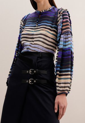 Femme portant un chemisier à manches longues rayé ondulé bleu et noir avec un col montant et une jupe noire dotée de deux brides avec boucles.