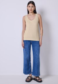 Beige tank top dengan leher V, dipadukan dengan celana jeans denim biru berpotongan lebar. Model mengenakan sandal hitam dengan aksen gesper.