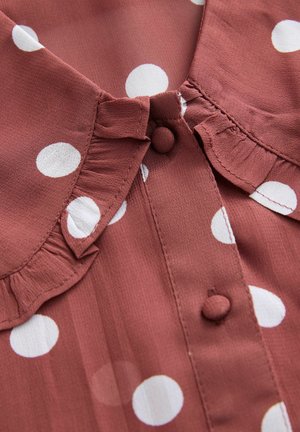 Rustfarvet bluse med hvide prikker, der har en flæset krave og knaplukning. Stoffet ser glat ud med en let tekstur.