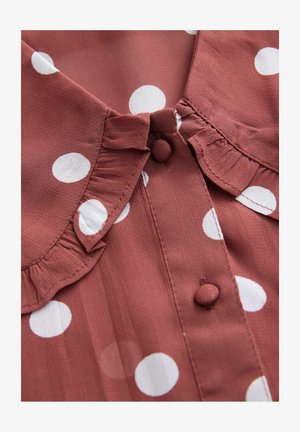 Rostfarbene Bluse mit weißen Punkten, ausgestattet mit einem gerüschten Kragen und Knopfverschluss. Der Stoff wirkt glatt und hat eine leichte Textur.
