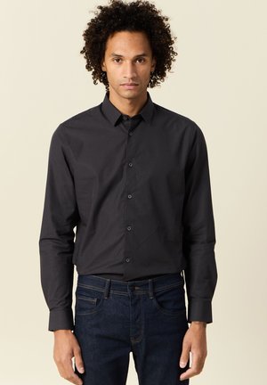 Camisa negra de botones con mangas largas, cuello y tapeta frontal. Tejido suave con una textura sutil; combinada con jeans de mezclilla azul oscuro.