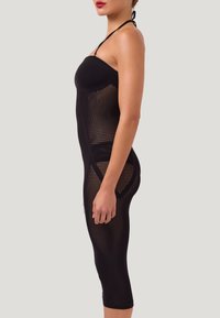 Donna di profilo che indossa un abito nero trasparente in rete con spalline ad anello che lascia intravedere la biancheria, su uno sfondo chiaro e uniforme.