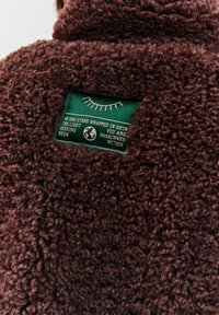 Hnědá texturovaná látka se zelenou náplatkou, na které je znázorněn ilustrace zavřeného oka a text: "Jsme hvězdy zabalené v kůži, světlo hledající tebe."