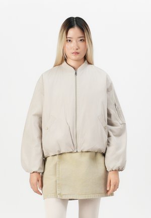 Jonge vrouw met steil half-gebleekt haar draagt een oversized beige bomberjack over een beige rok en witte leggings, staand tegen een witte achtergrond.