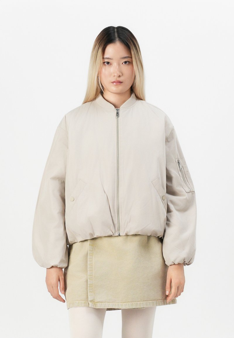 Mladá žena s rovnými poloblečenými vlasy na sobě má oversized béžovou bomber bundu přes béžovou sukni a bílé leginy, stojí na bílém pozadí.
