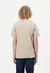 T-shirt en coton beige à manches courtes et col rond, vue de dos. La personne porte un jean bleu.