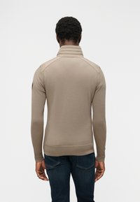 Άνδρας που φοράει μπεζ πλεκτό πουλόβερ με μακριά μανίκια και ψηλό γιακά, καθώς και σκούρο μπλε τζιν, στέκεται γυρισμένος με την πλάτη προς το απλό φόντο.