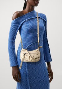 Žena v modrých žebrovaných pletených šatech s jedním ramínkem a malou světle béžovou crossbody kabelkou se metalovými nýty a zipem.