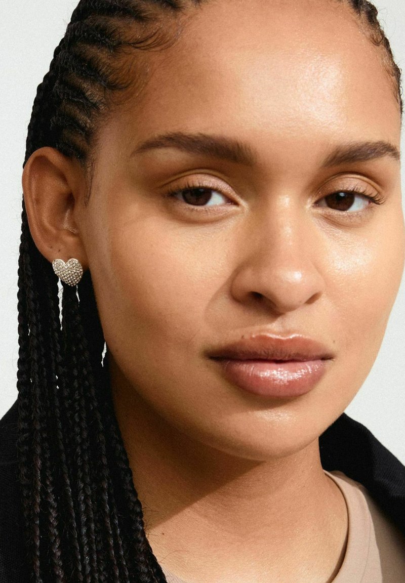 Pendientes en forma de corazón de plata con adornos texturizados, colgando de un stud, presentados en una modelo con pelo trenzado y maquillaje neutro.