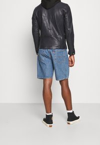 Svart läderjacka med synlig sömnad över en svart hoodie, parat med blå denimshorts och svarta högstövlade sneakers med vita snören.