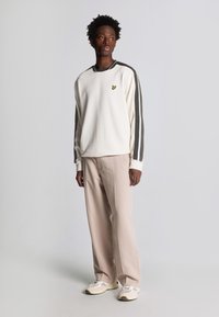 Felpa bianca con maniche a strisce grigie scure, piccolo logo giallo sul petto; abbinata a pantaloni beige chiaro e sneakers beige.