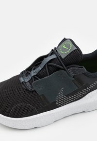 Black athletic shoe featuring a mesh upper, elastic laces, green accents, and a textured outsole with a speckled midsole.