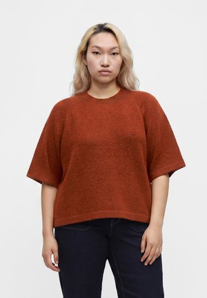 Ung person med langt blondt hår iført en løs rustfarvet kortærmet sweater og mørke bukser, stående mod en ensfarvet baggrund.