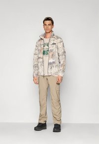 Kerge, mustriline halli värvusega lukuga jope, millel on pruunikaid toone; kombineeritud beežide t-särgi ja khaki värvi taskupükstega; mustad jalanõud.