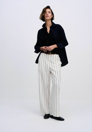 Nainen seisoo päällään löysä musta paita mustan topin päällä, valkoiset leveälahkeiset housut, joissa pystysuorat mustat raidat, musta vyö ja mustat kengät yksivärisellä taustalla.