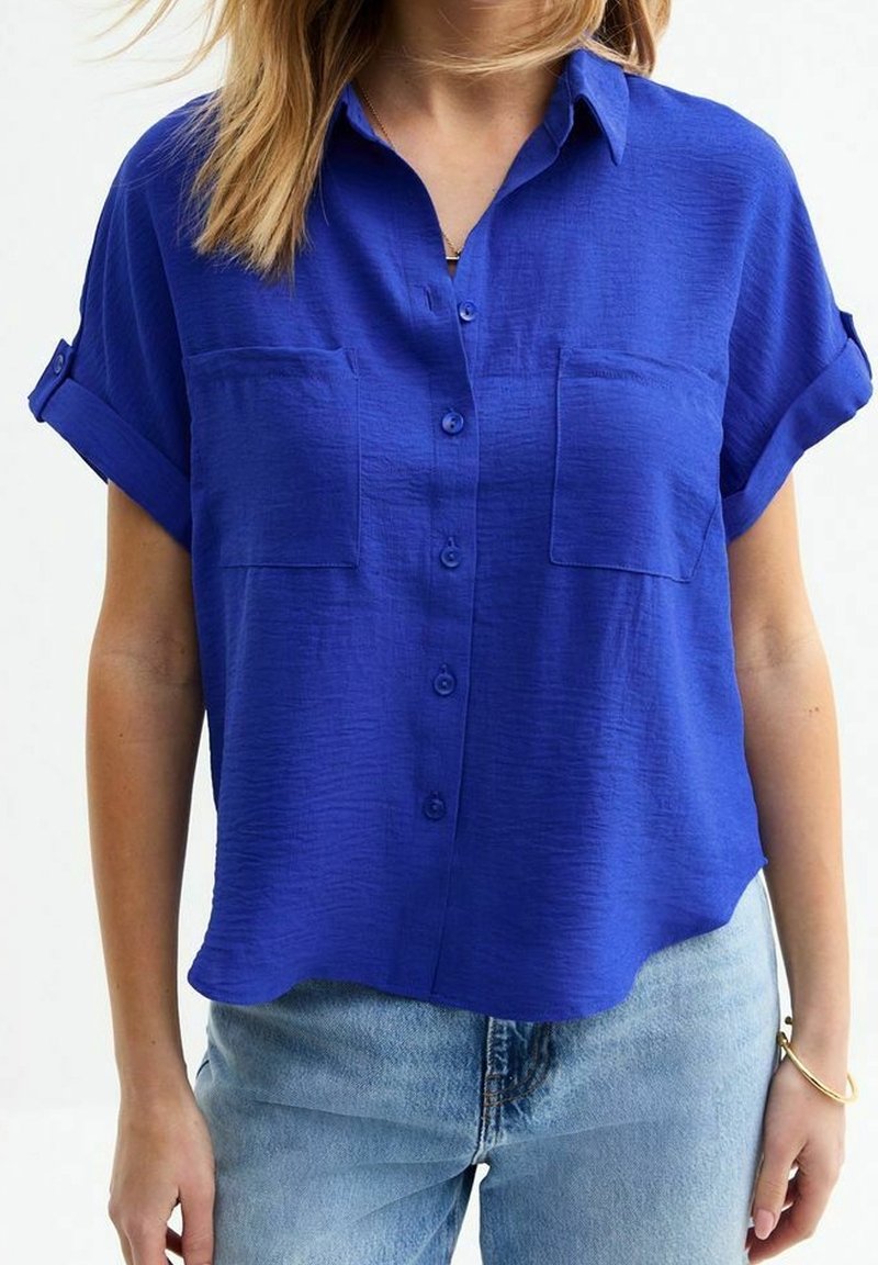 Femme portant une chemise bleu roi à manches courtes, boutonnée, avec deux poches sur le devant, et un jean bleu clair.