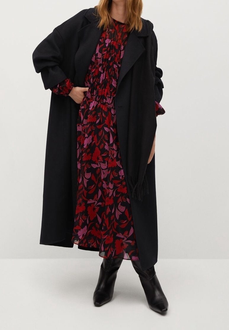 Femme portant un long manteau noir par-dessus une robe fleurie rouge et violette et des bottines noires, debout avec une main dans la poche.