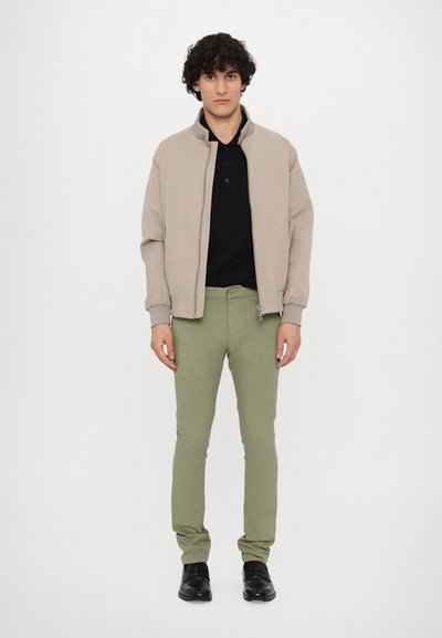 Ljusbeige zip-up jacka, svart polo-shirt, olivgröna slim-fit byxor och svarta skor; släta texturer med minimalistisk design.