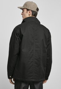 Svart jakke med en avslappet passform, med høy krage og kneppede mansjetter, laget av stoff med en glatt tekstur. Brukt med en beige caps.