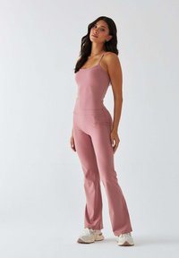 Jasnoróżowy strój sportowy z dopasowanym topem na ramiączkach i rozkloszowanymi spodniami, wykonany z gładkiej tkaniny, zestawiony z białymi trampkami.