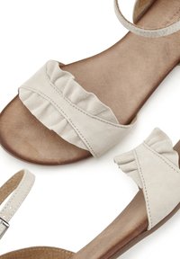 Beige mokkasandaalit, joissa on röyhelötyllin hihnat, avonainen kärki ja ruskea teksturoitu pohja yksinkertaisella nilkkahihnalla.