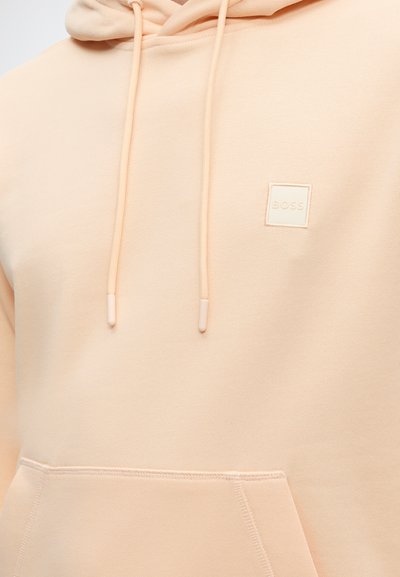 Sweat à capuche beige clair avec poche avant, cordons et petite étiquette carrée avec le logo "BOSS" sur la poitrine.