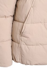 Close-up van een beige doorgestikte pufferjas met gestructureerde stof, horizontale stiksels en drukknoopsluitingen aan de voorkant.
