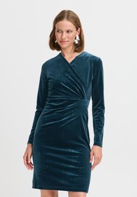 Teal sammetklänning med långa ärmar, V-ringning och knytning i midjan. Den släta texturen och den figurnära silhuetten framhäver figuren.