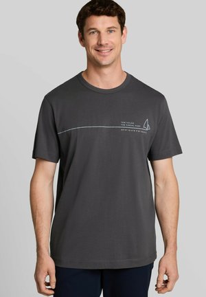 Mann mit kurzen braunen Haaren lächelt, trägt dunkelgraues T-Shirt mit dünner horizontaler Linie und kleinem Segelboot-Motiv auf der Brust.