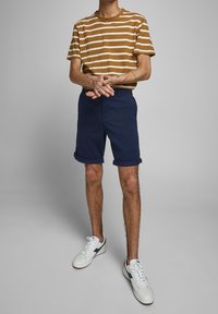 T-shirt a strisce marroni e bianche, pantaloni corti blu navy e sneakers bianche. I pantaloni corti sono arrotolati ai polsini; la t-shirt ha una vestibilità regolare. Abbigliamento casual.