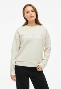 Mujer vestida con sudadera color crema con el texto "MINIMALISMO" y pantalones negros, de pie con una mano en el bolsillo frente a un fondo blanco.