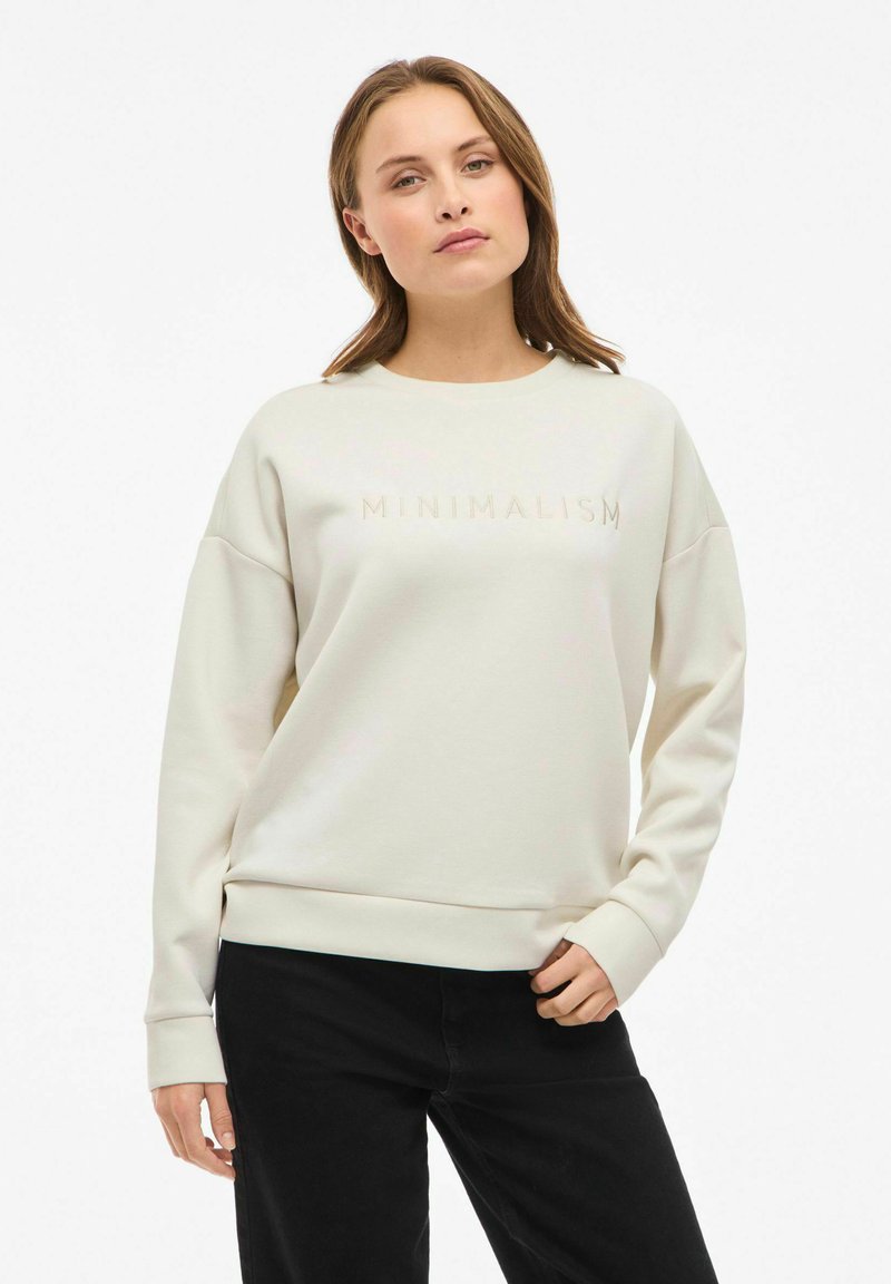 Mujer vestida con sudadera color crema con el texto "MINIMALISMO" y pantalones negros, de pie con una mano en el bolsillo frente a un fondo blanco.