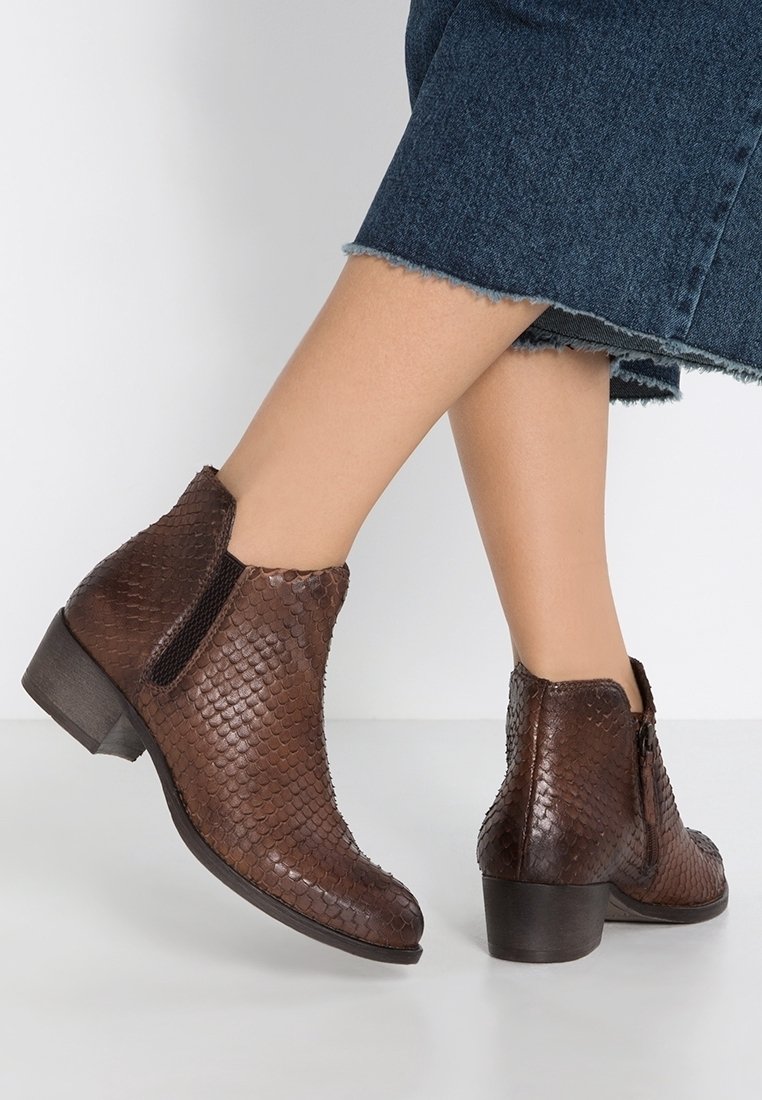 Bottes marron texturées à chevilles avec talons carrés portées avec un jean bleu court, montrées sur des jambes croisées sur un fond uni.