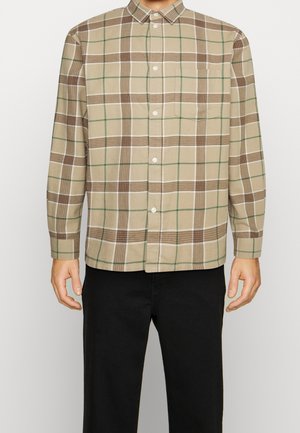 Homme portant une chemise beige à boutons avec un motif à carreaux marron et vert et un pantalon noir, debout les bras détendus le long du corps.