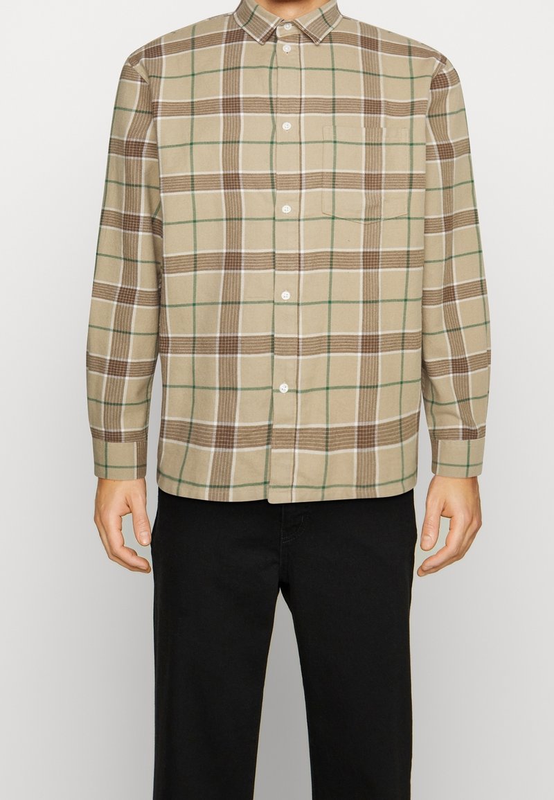 Homme portant une chemise beige à boutons avec un motif à carreaux marron et vert et un pantalon noir, debout les bras détendus le long du corps.