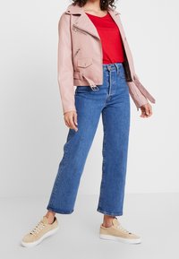 Giacca da motociclista in pelle rosa chiaro con accessori in argento, camicia rossa sotto, jeans blu a vita alta e sneakers beige.